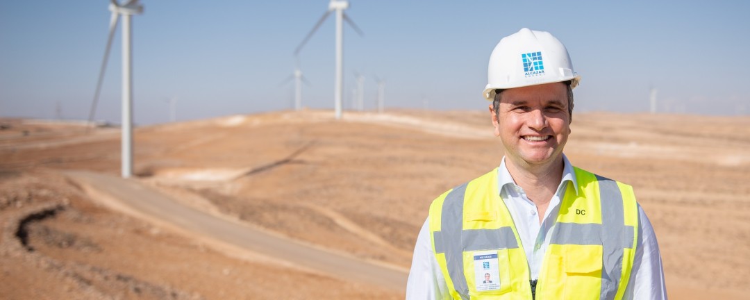 Mann mit gelber Warnweste steht in der Wüste. Im Hintergrund sind zwei Windräder zu sehen.
