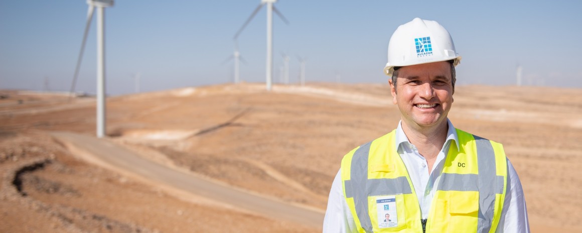 Mann mit gelber Warnweste steht in der Wüste. Im Hintergrund sind zwei Windräder zu sehen.