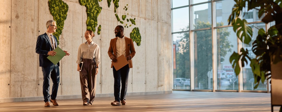 Drei Personen gehen mit Unterlagen durch eine moderne, lichtdurchflutete Lobby; an der Betonwand ist ein große Weltkarte aus Pflanzen angebracht.