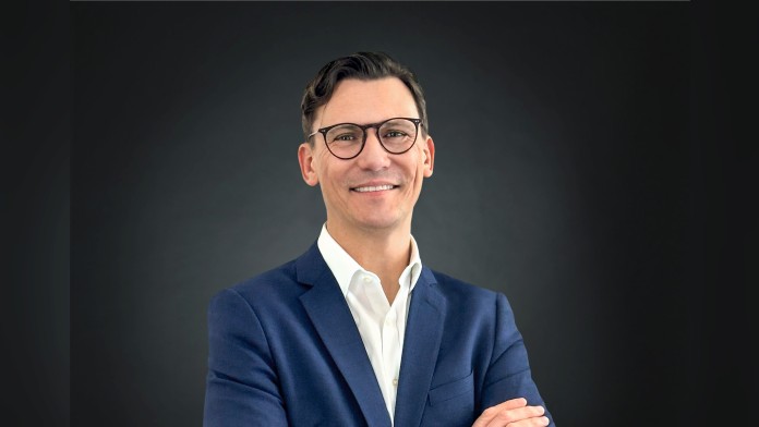 Portrait of a person in a blue suit with glasses, standing with folded arms against a dark gradient background.