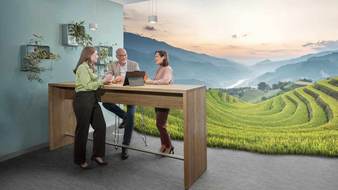 Drei Personen besprechen sich an einem Stehtisch mit Tablet vor einer großflächigen Darstellung grüner Terrassenfelder in einer Berglandschaft.