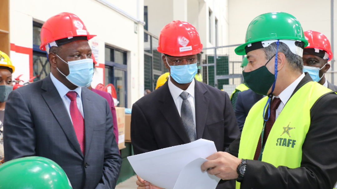 Gruppe von Personen mit Schutzhelmen und formeller Kleidung an einem Industrie- oder Baustellenstandort; eine Person mit „STAFF“-Weste zeigt Dokumente