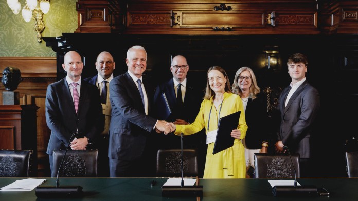 Several people in business attire stand behind a table in a historic room while two individuals shake hands and hold folders.