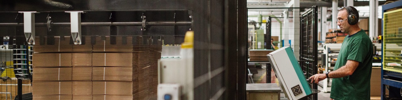 Arbeiter bedient Steuerpult in einer Fabrik; im Vordergrund liegt ein Stapel flacher Kartonbögen auf einer Palette.