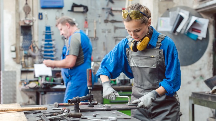 Werkstatt mit zwei Personen bei der Metallbearbeitung, umgeben von Werkzeugen und Maschinen.