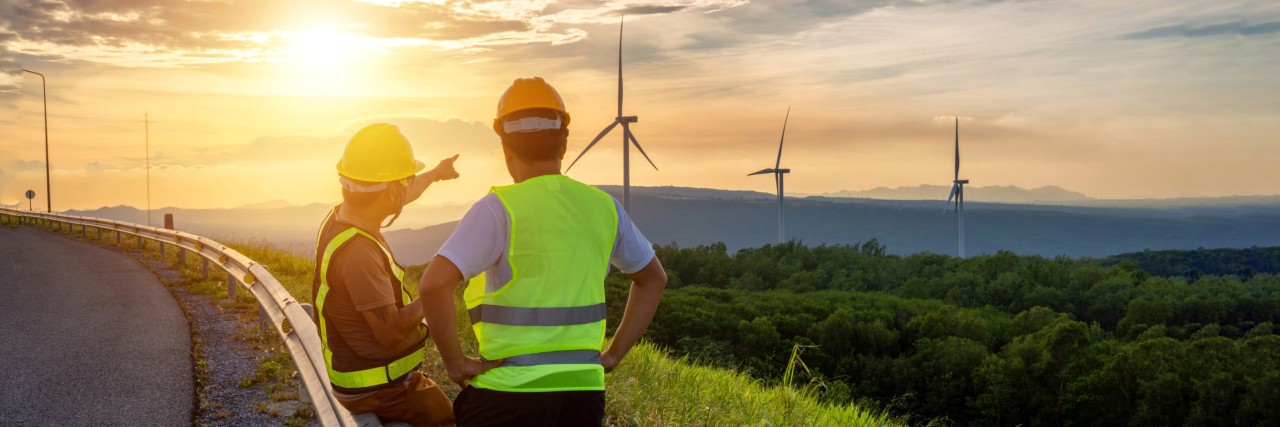 Zwei Personen in Warnwesten stehen an einer Straße und blicken auf eine Landschaft mit mehreren Windkraftanlagen bei Sonnenuntergang.